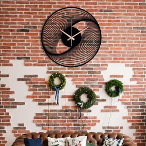 May include: Black, circular wall clock with a unique, layered design. The clock hands are light brown, and the clock is mounted on a red brick wall. The clock's design creates a spiral effect. Below the clock are three green wreaths.