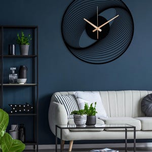 May include: A modern black wall clock with a unique, abstract design. The clock features a circular shape with a swirling pattern and gold-colored hands. It is mounted on a dark blue wall, complementing the contemporary interior decor.