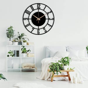 A black metal wall clock with Roman numeral markings and a black face. The clock is hanging on a white wall above a white bed with white bedding and green plants.