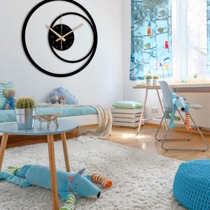Pu&ograve; includere: Un moderno orologio da parete nero dal design unico. L'orologio presenta un quadrante circolare con un cerchio interno pi&ugrave; piccolo e lancette di colore chiaro. L'orologio &egrave; montato su una parete bianca nella camera da letto di un bambino, con uno schema di colori blu e bianco.