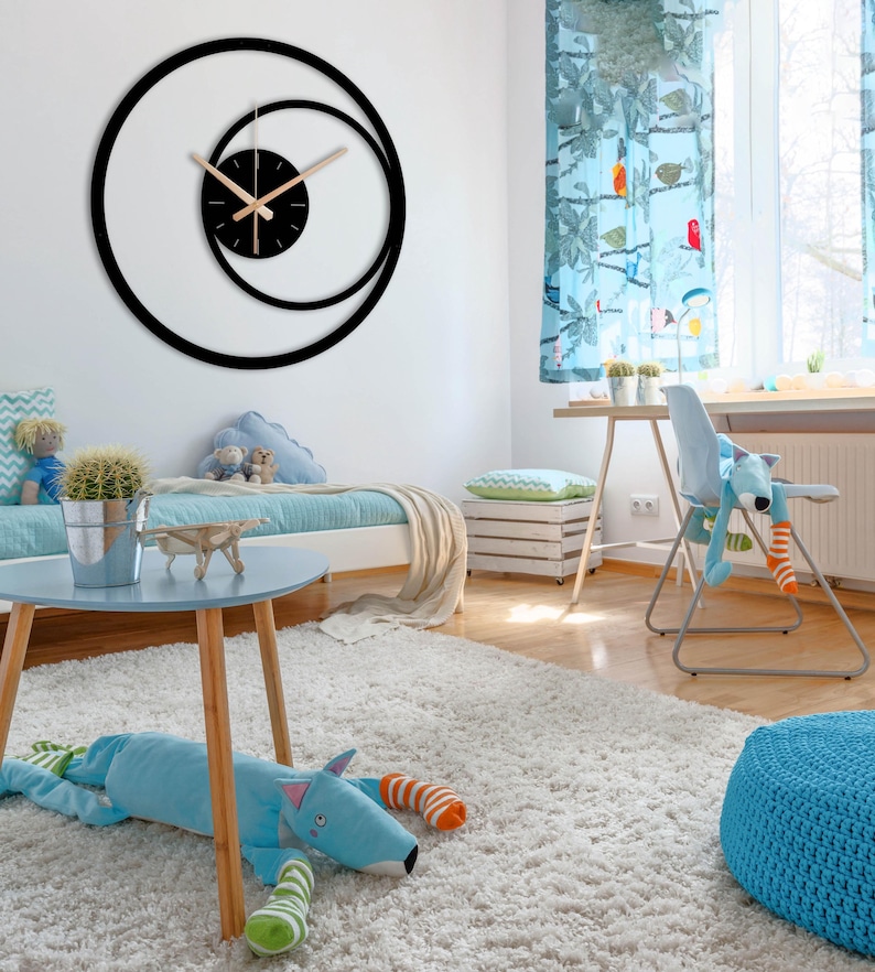 Pu&ograve; includere: Un moderno orologio da parete nero dal design unico. L'orologio presenta un quadrante circolare con un cerchio interno pi&ugrave; piccolo e lancette di colore chiaro. L'orologio &egrave; montato su una parete bianca nella camera da letto di un bambino, con uno schema di colori blu e bianco.