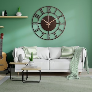 K&ouml;nnte beinhalten: Eine braune Wanduhr aus Metall mit r&ouml;mischen Ziffern und einem dekorativen Au&szlig;enring. Die Uhr h&auml;ngt an einer gr&uuml;nen Wand &uuml;ber einem wei&szlig;en Sofa mit gr&uuml;n-wei&szlig; gestreiften Kissen.