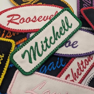 May include: A collection of embroidered name patches in various colors and shapes. The patches feature names like "Mitchell" and "Roosevelt" in different fonts and colors, with contrasting border stitching. The patches are on a white background.