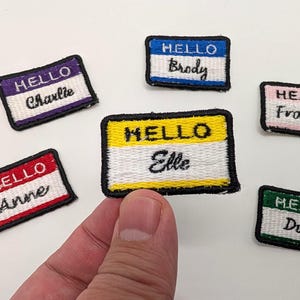 May include: Five colorful embroidered iron-on patches with the text "Hello" and a name on each patch. The names are Brody, Charlie, Elle, Frank, and Duke.
