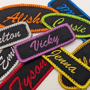 May include: A collection of embroidered name patches in various colors. Each patch features a name in a cursive font, with different border colors like orange, green, blue, and yellow. The patches are on a black background.