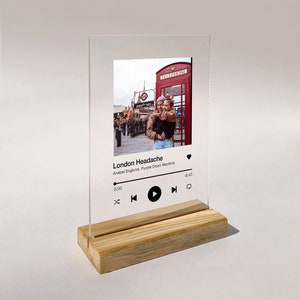 May include: A clear acrylic music player stand with a wooden base. The stand features a photo of two people in front of a red telephone box. The music player display shows the song "London Headache" by Anabel Englund, Purple Disco Machine.