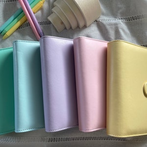 Könnte beinhalten: Fünf pastellfarbene, ledergebundene Notizbücher mit Klappenverschluss. Die Notizbücher sind in einer Reihe von links nach rechts angeordnet: hellgrün, blau, lavendel, rosa und gelb.