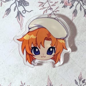 May include: A clear acrylic charm featuring a cartoon character with orange hair and a white beret. The character has large blue eyes and a small smile. The charm is set against a floral background with pink and purple flowers.