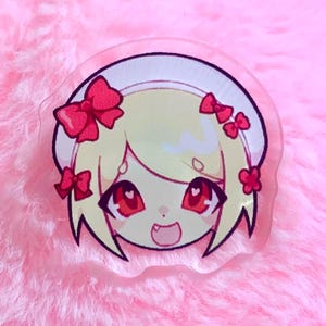 Könnte beinhalten: Ein klarer Acryl-Anhänger mit einem Anime-Charakter mit blonden Haaren, roten Augen und roten Schleifen. Der Charakter hat einen lächelnden Ausdruck. Der Anhänger ist vor einem rosa, flauschigen Hintergrund platziert.