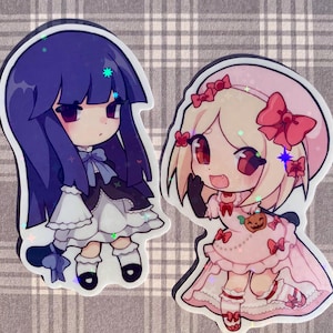 Puede incluir: Dos pegatinas de estilo anime. Una presenta un personaje con pelo largo morado, un vestido blanco y negro y un lazo azul. La otra tiene pelo rubio, un vestido rosa y una calabaza. Ambas tienen detalles brillantes.