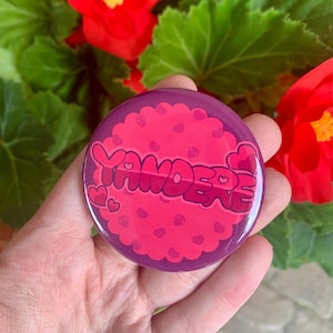 May include: A purple and pink button with the word "Yandere" in a pink, bubbly font. The button is covered in small pink hearts and has a glossy finish.