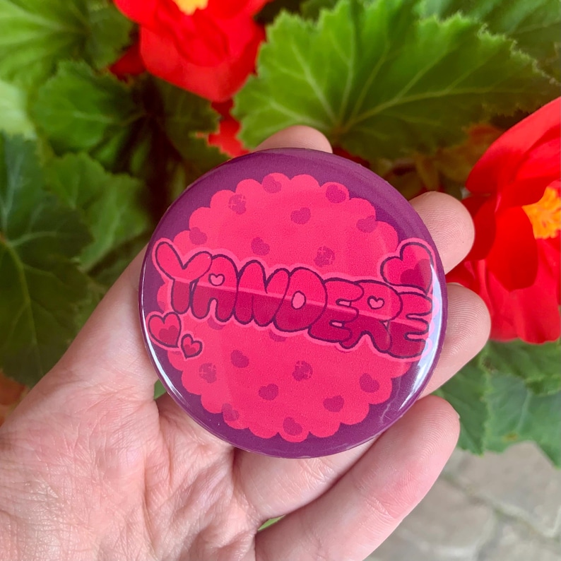 May include: A purple and pink button with the word "Yandere" in a pink, bubbly font. The button is covered in small pink hearts and has a glossy finish.