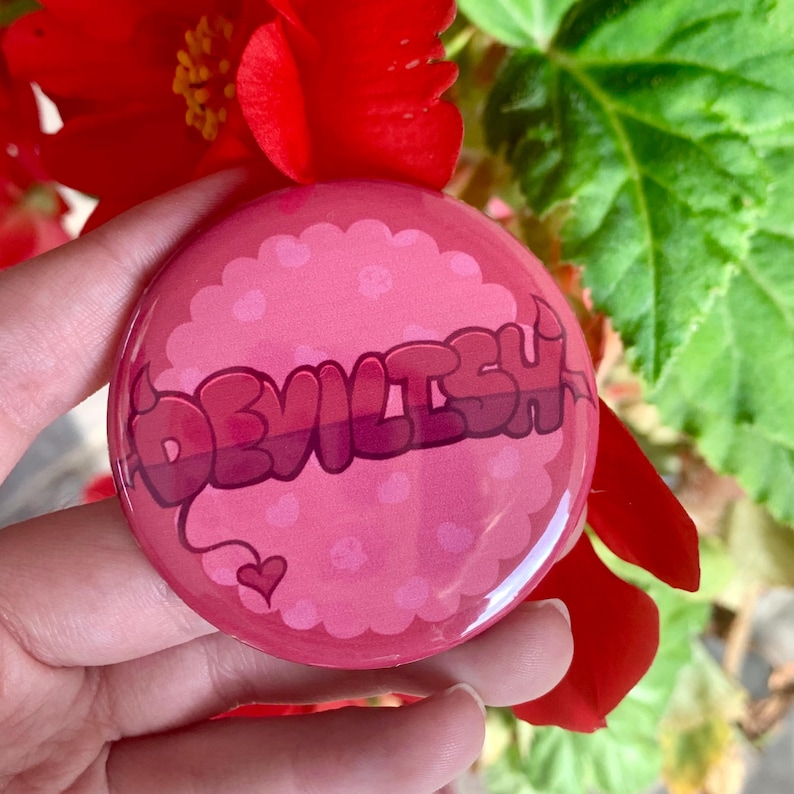 May include: Pink button with a white outline and a pink heart. The button says "DEVILISH" in pink and white letters with a small devil tail and horns.