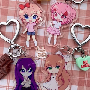 May include: Four clear acrylic keychains featuring chibi-style anime characters with colorful hair and outfits. Each keychain has a silver heart-shaped clasp and a small charm, including an ice cream cone, a chocolate bar, and a green heart with text.