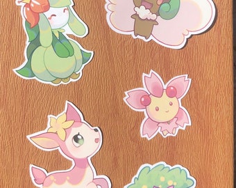 Könnte beinhalten: Fünf bunte Comic-Aufkleber mit verschiedenen Pokémon-Charakteren. Die Aufkleber befinden sich auf einem braunen Hintergrund. Die Charaktere umfassen ein rosa Reh, eine grün-rosa Blume, eine grün-weiße Blume, eine braun-rosa Wolke und einen grün-weißen Igel.