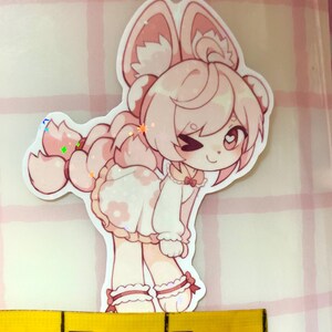 May include: A pink and white sticker of a cartoon character with large ears and a dress. The sticker is about 5 cm in height, with a glittery finish. The character is winking. The sticker is on a pink and white checkered background.
