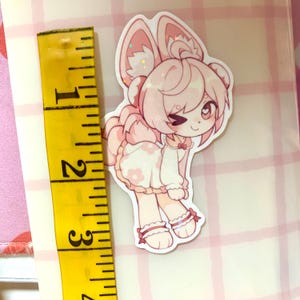 May include: A pink and white sticker of a cartoon character with bunny ears and a dress with flower details. The sticker is approximately 6.35 cm tall. The character is winking and has a small bow. The sticker has glitter accents.
