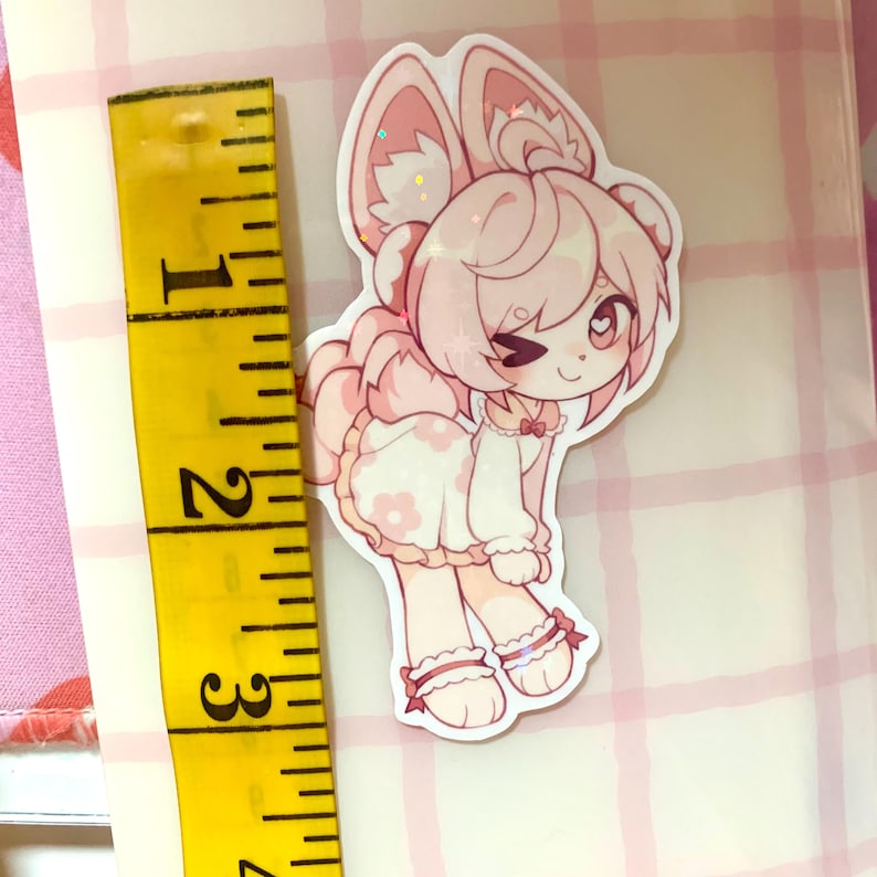 May include: A pink and white sticker of a cartoon character with bunny ears and a dress with flower details. The sticker is approximately 6.35 cm tall. The character is winking and has a small bow. The sticker has glitter accents.