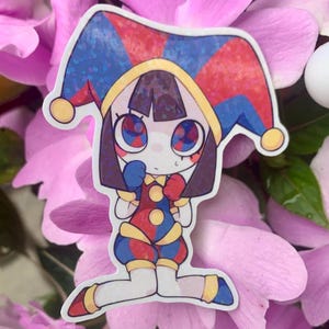 Peut inclure: Autocollant coloré représentant un personnage de bouffon de dessin animé avec une tenue et un chapeau rouges, bleus et jaunes. Le bouffon a de grands yeux et est à genoux. L'autocollant est posé sur un fond de fleurs roses.