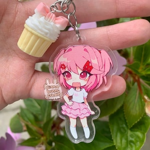 May include: An acrylic keychain featuring a pink-haired anime character with a red hair clip, wearing a white top and pink ruffled skirt, paired with a small cupcake charm with a pink bow. The keychain includes the text cinna bunny cream etsy.com.
