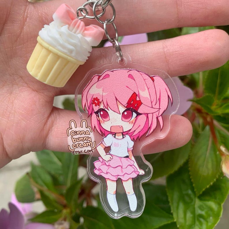 May include: An acrylic keychain featuring a pink-haired anime character with a red hair clip, wearing a white top and pink ruffled skirt, paired with a small cupcake charm with a pink bow. The keychain includes the text cinna bunny cream etsy.com.