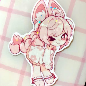May include: A pink and white sticker featuring a cartoon character with large ears, a winking eye, and a dress with flower details. The sticker has a holographic, glittery effect, and is set against a pink and white checkered background.