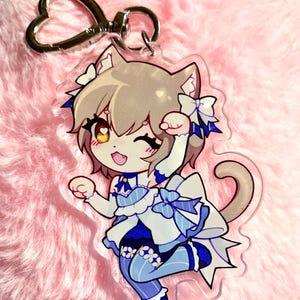 Puede incluir: Un llavero acrílico transparente con un personaje de dibujos animados con orejas de gato, vestido con un atuendo azul y blanco con lazos. Tiene un cierre plateado en forma de corazón. El fondo es un material suave, rosa y difuso.