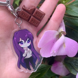 May include: A keychain featuring a clear acrylic charm of an anime-style character with long purple hair wearing a white sweater and black pants, paired with a small brown chocolate bar charm embossed with the word MILK on each square segment.
