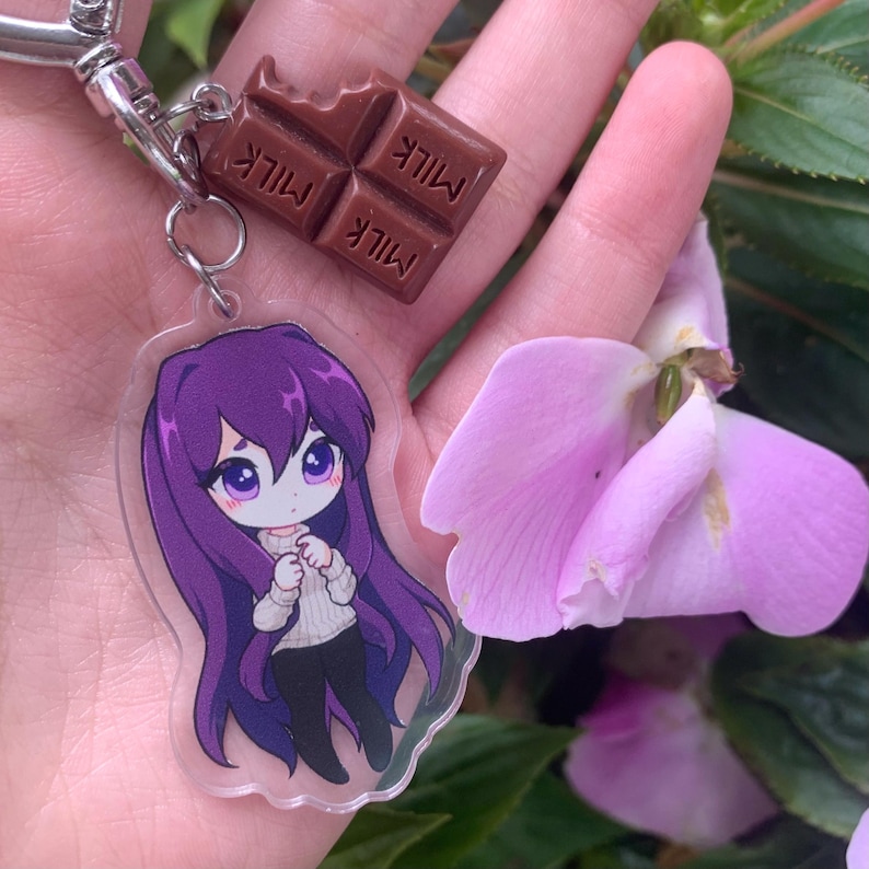 May include: A keychain featuring a clear acrylic charm of an anime-style character with long purple hair wearing a white sweater and black pants, paired with a small brown chocolate bar charm embossed with the word MILK on each square segment.