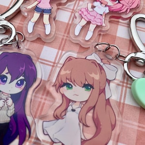 May include: Four acrylic keychains featuring chibi-style anime characters. One character has peach hair, one has pink hair with a red bow, one has long purple hair in a cream sweater, and one has long brown hair with a white bow, all with silver clasps.