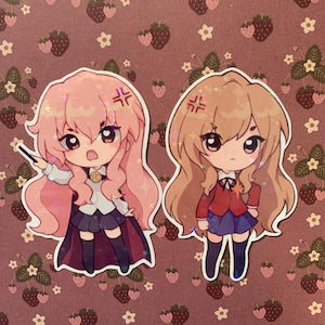 May include: Two anime character stickers with pink and brown hair. The characters are both girls with large eyes and are wearing school uniforms. The stickers are on a pink and white background with a strawberry pattern.