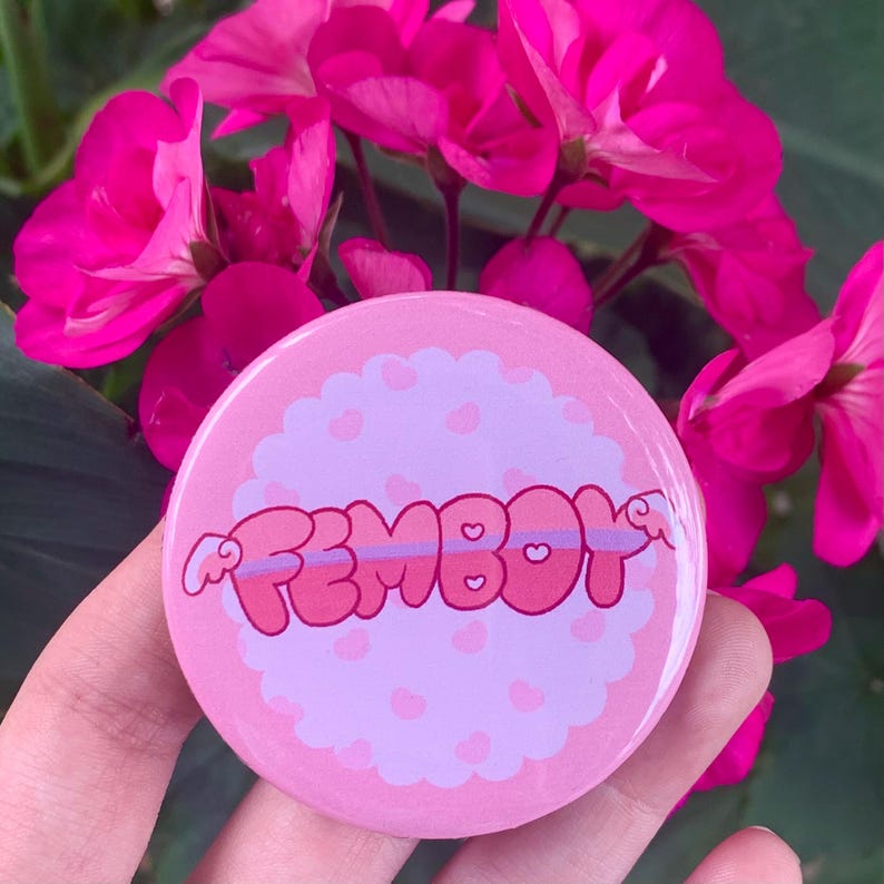 May include: Pink and purple button with the word "FEMBOY" in bubble letters with pink and purple hearts around it.