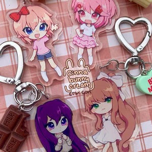 May include: Four acrylic keychains featuring anime-style characters with colorful hair and outfits. The keychains include metal heart-shaped clasps and small charms shaped like ice cream, cupcakes, and chocolate bars. Text reads Cinna bunny cream ETSY. COM.