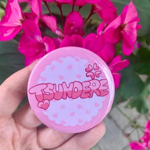 May include: Pink and white button with the word "Tsundere" in pink and white lettering. The button has a pink and white heart design.