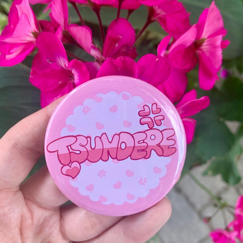 May include: Pink and white button with the word "Tsundere" in pink and white lettering. The button has a pink and white heart design.