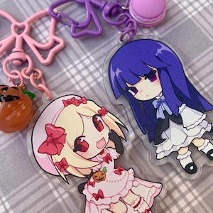 Può includere: Una collezione di portachiavi in stile anime con personaggi dei cartoni animati. Un portachiavi presenta un personaggio biondo in un vestito rosa con fiocchi rossi e una zucca. Un altro portachiavi mostra un personaggio con i capelli viola e un vestito bianco. Altri portachiavi includono un fiocco rosa, un fiocco viola e un macaron rosa.