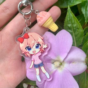 May include: A clear acrylic keychain featuring a cartoon character with pink hair, a red bow, a pink shirt, and blue shorts, attached to a silver metal ring with a small, textured pink ice cream cone charm with a yellow waffle cone base.