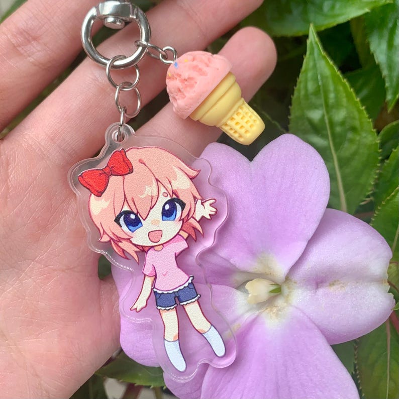 May include: A clear acrylic keychain featuring a cartoon character with pink hair, a red bow, a pink shirt, and blue shorts, attached to a silver metal ring with a small, textured pink ice cream cone charm with a yellow waffle cone base.