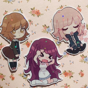 Op de afbeelding: Drie cartoon-personage stickers met glitteraccenten. De personages zijn een meisje met bruin haar en een zwart-witte outfit, een meisje met roze haar en een zwart-witte outfit en een meisje met paars haar en een wit-roze outfit.