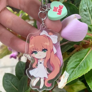 May include: A clear acrylic keychain featuring a chibi-style anime character with long peach-colored hair, a white bow, and a white dress. Attached to the silver metal ring is a small, heart-shaped mint green charm with the red text BE MINE.