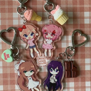 May include: Four acrylic keychains featuring chibi-style anime characters with heart-shaped clasps. Charms include a light pink ice cream cone, a white cupcake, a green heart with the text BE MINE, and a brown chocolate bar. The characters have distinct hair colors including light brown, pink, brown, and purple.