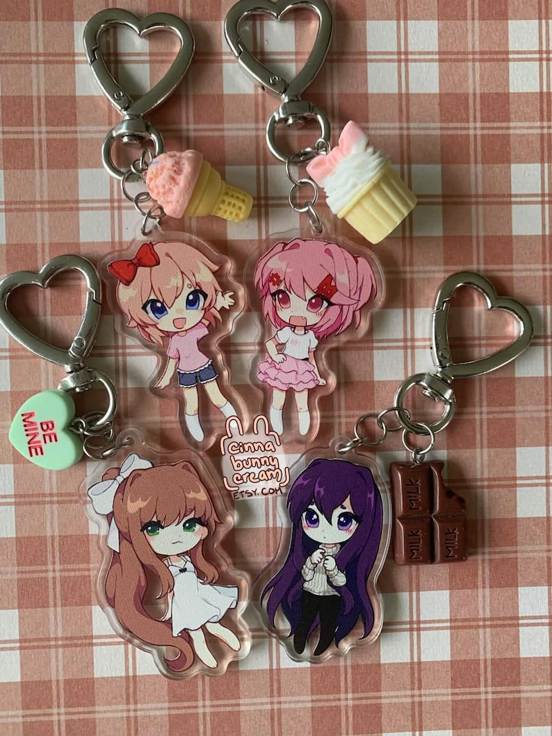 May include: Four acrylic keychains featuring chibi-style anime characters with heart-shaped clasps. Charms include a light pink ice cream cone, a white cupcake, a green heart with the text BE MINE, and a brown chocolate bar. The characters have distinct hair colors including light brown, pink, brown, and purple.