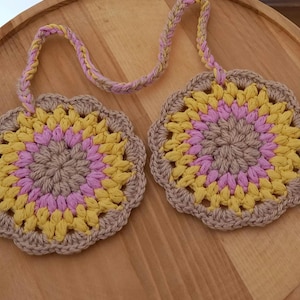 May include: Two crocheted coasters with a brown, pink, and yellow color pattern. The coasters are connected by a crocheted string.