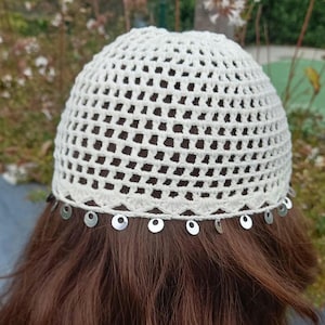 May include: A cream-colored crochet hat with a circular open weave design. The hat is adorned with small, silver-colored circular embellishments along the bottom edge. The hat is worn on brown hair.