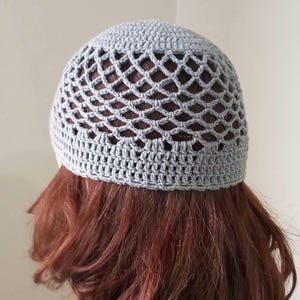 May include: A light gray crocheted beanie hat with an open, net-like design. The hat is handmade and appears to be made of a soft yarn. The hat is shown from the back, with the wearer's red hair visible through the open weave.