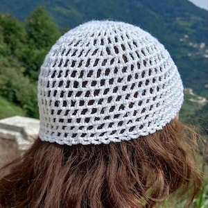 May include: A white, open-weave crochet hat with subtle sequin accents. The hat is a light, airy design, perfect for warm weather. The hat is shown from the back, with brown hair visible beneath it. The background features a blurred view of green hills and a blue sky.