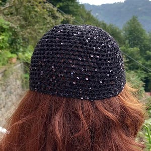 May include: A black crocheted skull cap with small, shiny sequins. The cap is worn on a head with long, auburn hair. The background features green trees and a mountain range.
