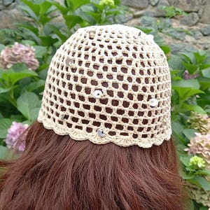May include: A crocheted hat in a light beige color with a net-like pattern. The hat is adorned with small, silver-colored sequins and has a scalloped edge. The hat is shown from the back, with brown hair visible beneath.