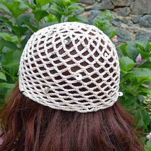 May include: A cream-colored crocheted hat with a net-like design, adorned with small, silver sequins. The hat is worn on brown hair, with a natural green and stone background.