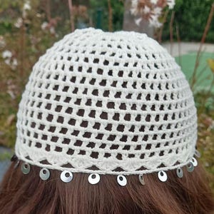 May include: A cream-colored, crocheted skull cap with a delicate openwork design. The cap is adorned with small, silver-colored circular embellishments along the bottom edge. The cap is shown on a person with brown hair.